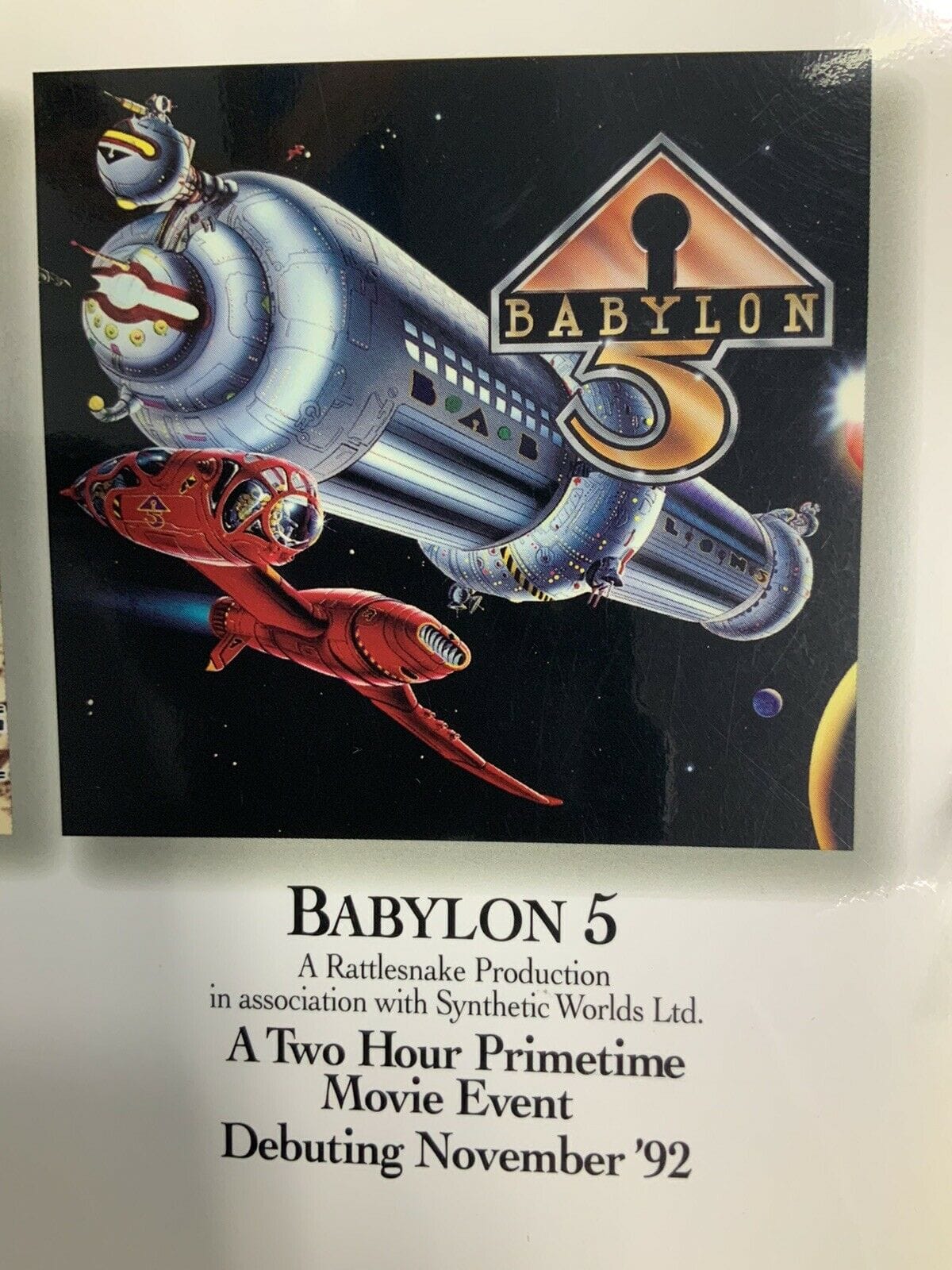 In this promotional image, BABYLON 5 is more like a silver tube with a docking port maw and little greeblies everywhere. A red ship, that resembles a sail boat that's capsized, whooshes past the station. A glimmer gold triangle with keyhole BABYLON 5 logo is in the upper right corner.